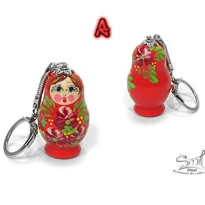 May include: Two red Matryoshka doll keychains. The wooden dolls are painted with floral designs in green, pink, and white. Each doll has a silver keychain attached. The text "A" is above the dolls.