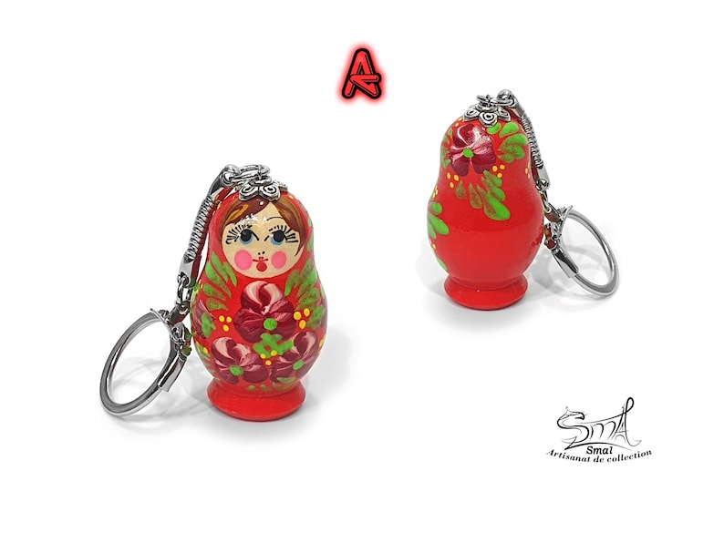 May include: Two red Matryoshka doll keychains. The wooden dolls are painted with floral designs in green, pink, and white. Each doll has a silver keychain attached. The text "A" is above the dolls.