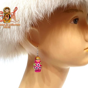 May include: A pair of dangle earrings featuring a Russian nesting doll design. The earrings are painted in shades of pink, green, and white, with a silver-colored hook. The earrings are a unique accessory.
