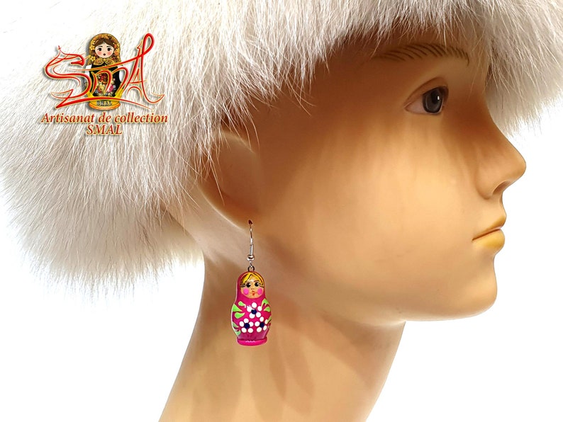 May include: A pair of dangle earrings featuring a Russian nesting doll design. The earrings are painted in shades of pink, green, and white, with a silver-colored hook. The earrings are a unique accessory.