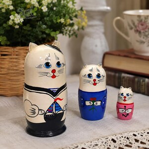 Three hand-painted cat sailor matryoshka dolls displayed on a cozy linen tablecloth, with a flower basket, decorative vase, teacup and vintage books in the background. The set includes a large black sailor cat, a blue-dressed cat and a small pink sailor cat