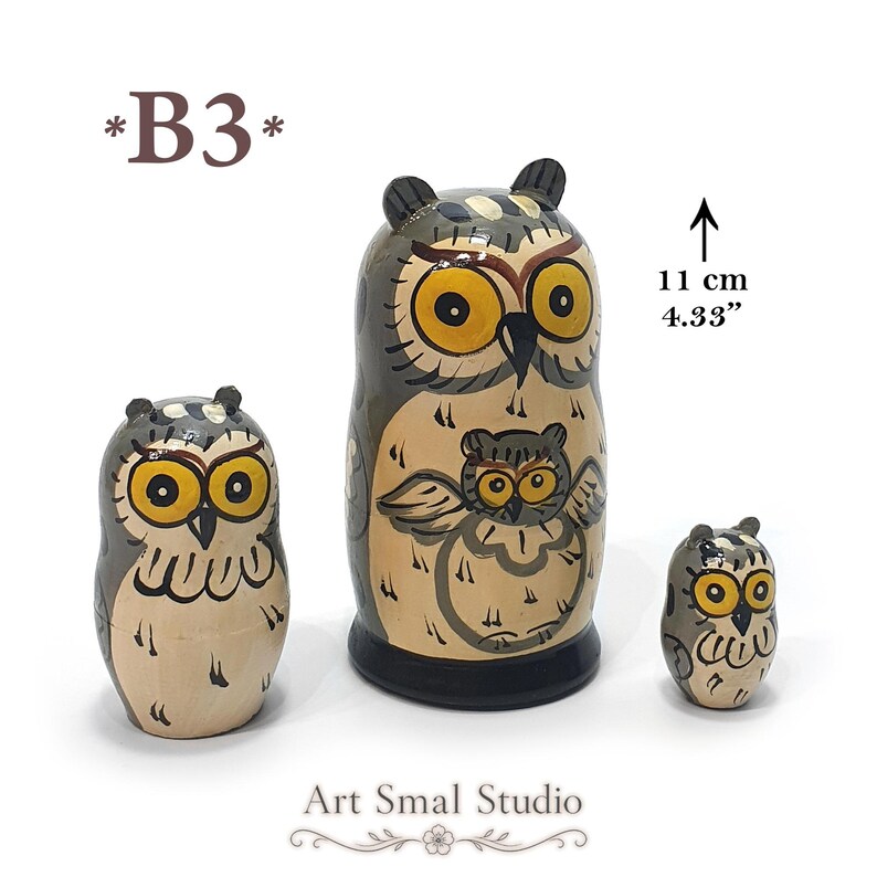 Three-piece hand-painted owl matryoshka nesting doll set in gray and cream, featuring expressive yellow eyes and detailed folk-art designs; largest doll measures 11 cm (4.33 inches) tall. Modèle B3
