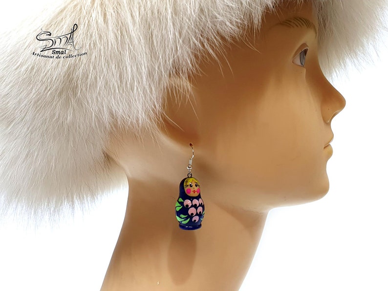 May include: A single Matryoshka doll earring. The earring is dark blue with a pink and green floral design. The doll has a yellow head and a pink face. The earring is hanging from a silver hook.