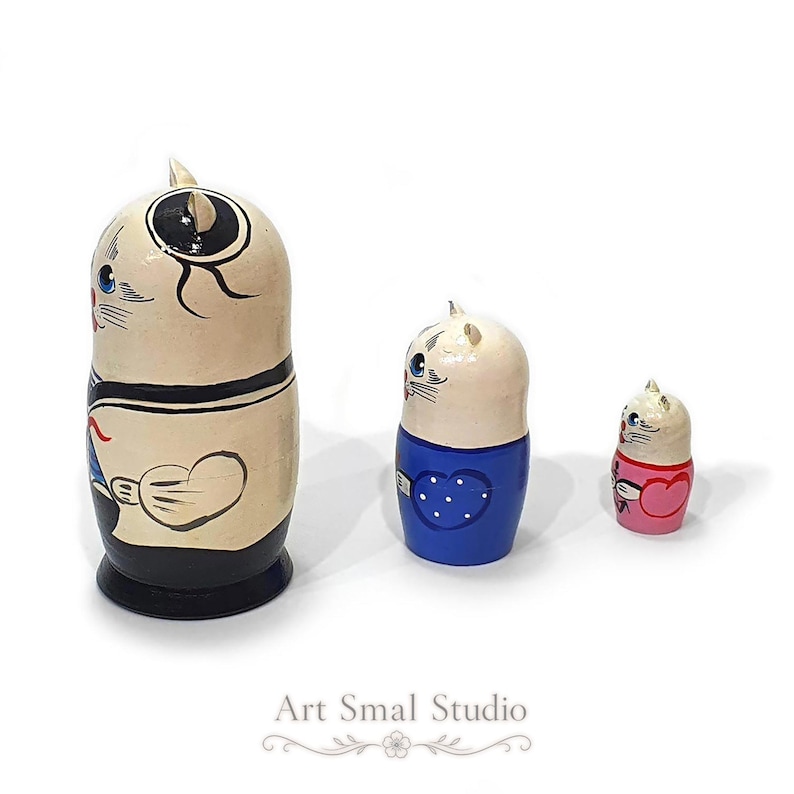 A side view highlights the hand-painted cat sailor matryoshka set, showing the nautical costumes, heart motifs and blue-and-pink details on the three wooden nesting dolls against a white background