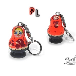 May include: A red and black wooden keychain shaped like a traditional Russian nesting doll, or Matryoshka doll. The doll has a painted face with blue eyes and a red and white patterned dress. The keychain has a silver metal ring.