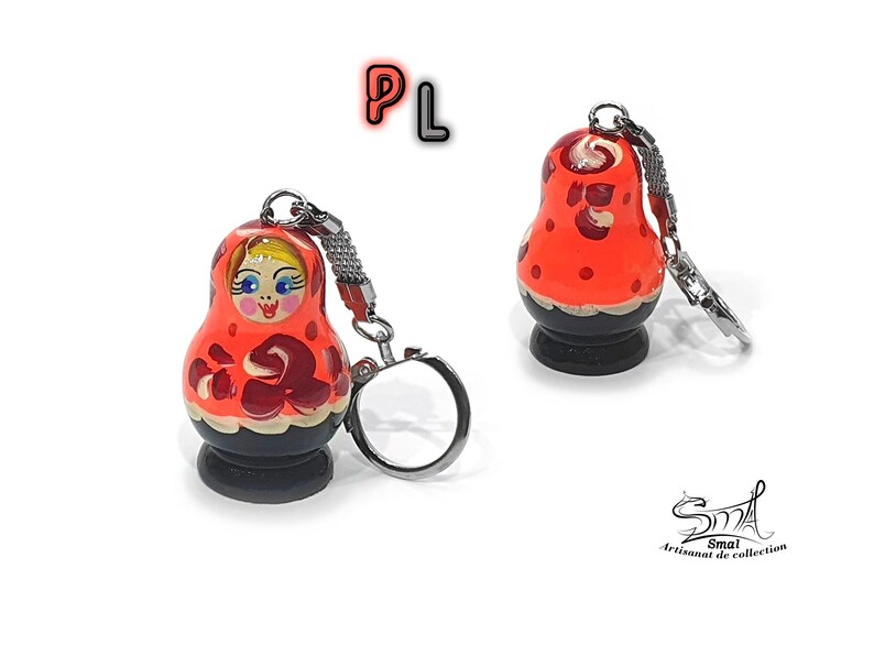 May include: A red and black wooden keychain shaped like a traditional Russian nesting doll, or Matryoshka doll. The doll has a painted face with blue eyes and a red and white patterned dress. The keychain has a silver metal ring.