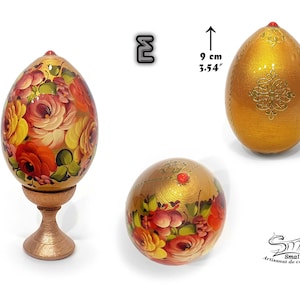 Œuf de Pâques or lumineux au fini nacré, décor floral peint à la main dans l’esprit de l’art russe, enrichi de motifs dorés et d’un cabochon rouge, présenté sur socle, hauteur totale 12,5 cm, hauteur de l’œuf 9 cm