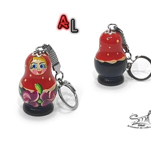 May include: A red and black wooden keychain shaped like a traditional Russian nesting doll, or matryoshka doll. The doll has a painted face with blue eyes and a red and white patterned dress. The keychain has a silver metal ring.