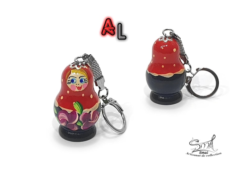 May include: A red and black wooden keychain shaped like a traditional Russian nesting doll, or matryoshka doll. The doll has a painted face with blue eyes and a red and white patterned dress. The keychain has a silver metal ring.