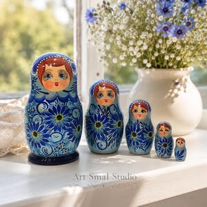Matriochka russe bleue exposée sur un rebord de fenêtre baigné de lumière, avec un bouquet de fleurs en arrière-plan. Ensemble de 5 poupées gigognes en bois peintes à la main, parfaites pour une décoration intérieure élégante et authentique.