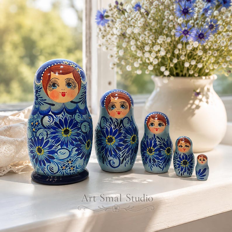 Matriochka russe bleue exposée sur un rebord de fenêtre baigné de lumière, avec un bouquet de fleurs en arrière-plan. Ensemble de 5 poupées gigognes en bois peintes à la main, parfaites pour une décoration intérieure élégante et authentique.