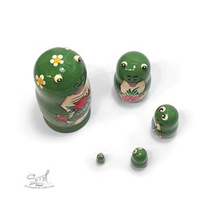 From an elevated angle, five green frog nesting dolls are arranged separately on a white background, revealing their rounded shapes, glossy finish, raised eyes and floral frog-themed artwork