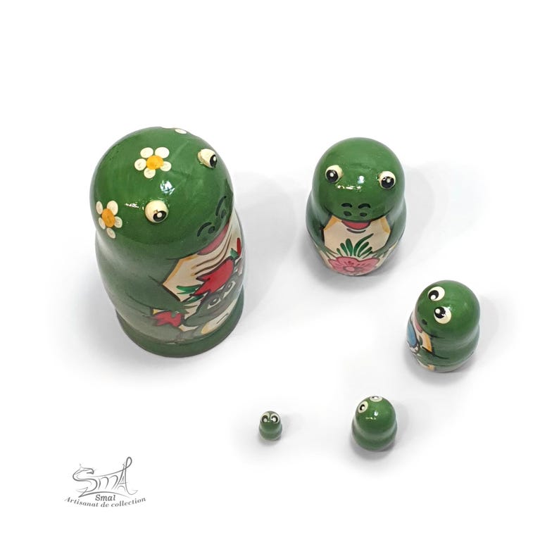 From an elevated angle, five green frog nesting dolls are arranged separately on a white background, revealing their rounded shapes, glossy finish, raised eyes and floral frog-themed artwork