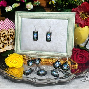 May include: A pair of black and blue earrings with floral designs. The earrings are displayed on a silver tray with other earrings and a yellow rose.