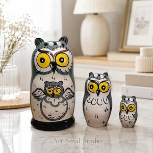 Hand-painted Russian owl nesting dolls in gray and cream, three-piece matryoshka set with bright yellow eyes displayed on a light marble surface in a cozy modern home interior.