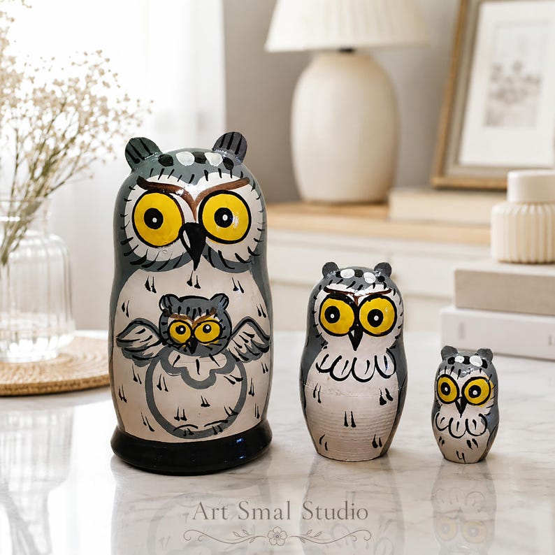 Hand-painted Russian owl nesting dolls in gray and cream, three-piece matryoshka set with bright yellow eyes displayed on a light marble surface in a cozy modern home interior.