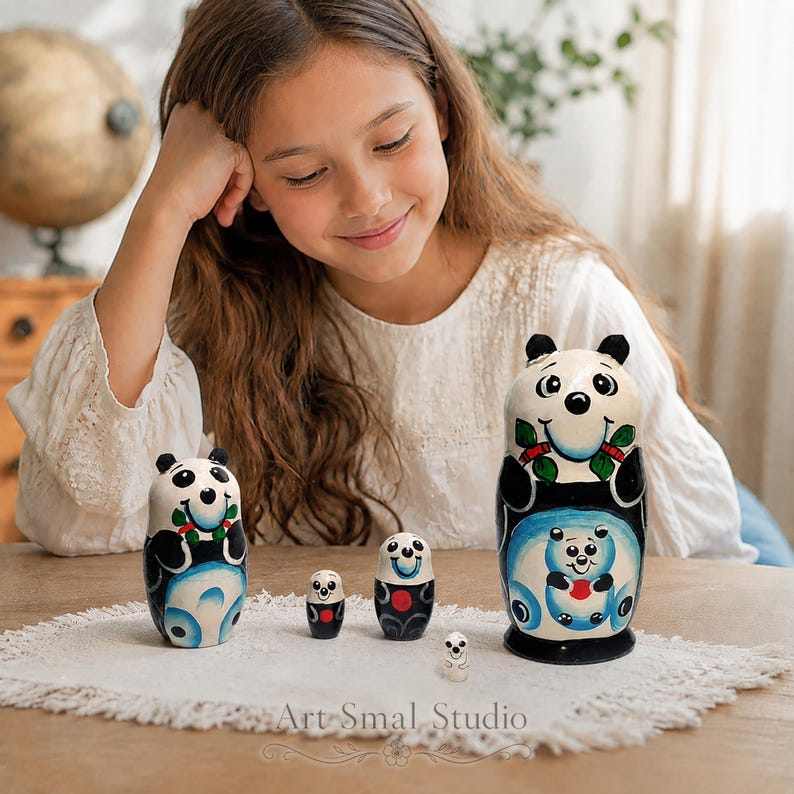 Hand-painted panda matryoshka nesting dolls, a set of five Russian wooden dolls displayed on a white fringed cloth, with a young girl admiring them in a warm home interior. Colorful panda designs feature black, white, blue, green and red details