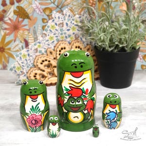 A colorful frog nesting doll collection is arranged on a tabletop, surrounded by floral textiles, handmade wooden decorations and a potted plant, highlighting the cheerful hand-painted frog artwork