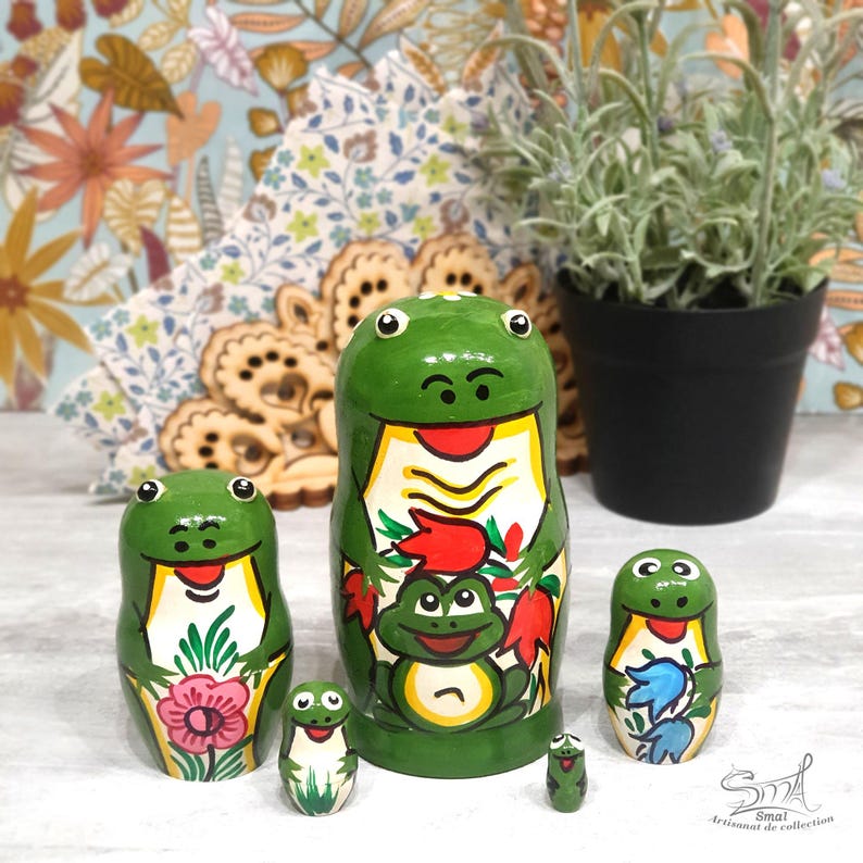 A colorful frog nesting doll collection is arranged on a tabletop, surrounded by floral textiles, handmade wooden decorations and a potted plant, highlighting the cheerful hand-painted frog artwork