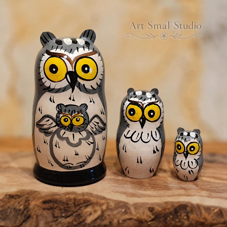 Three-piece owl matryoshka nesting doll set with expressive yellow eyes, hand-painted gray and cream folk-art details, displayed on a rustic wooden surface against a softly blurred warm beige background