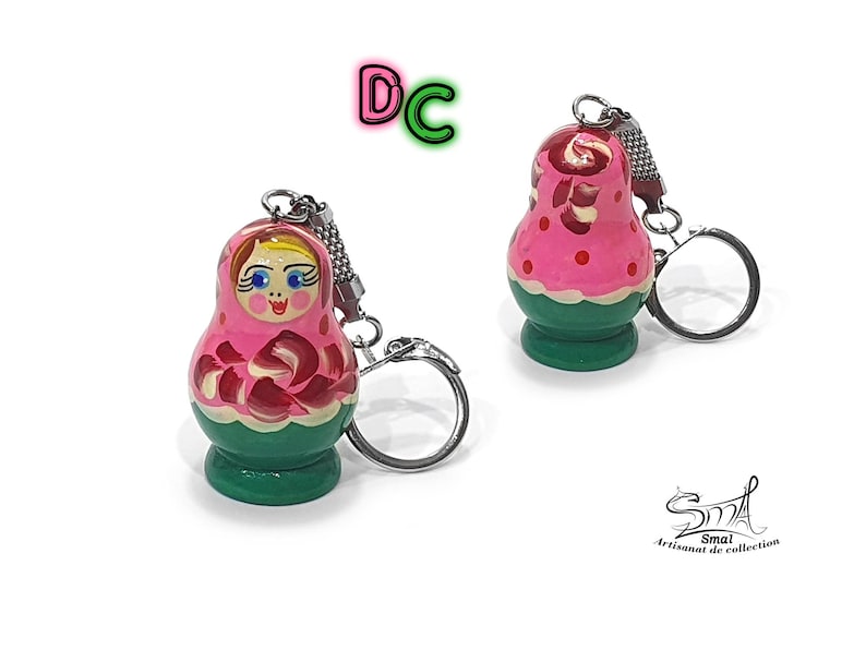 May include: A pink and green wooden nesting doll keychain with a painted face and floral design. The keychain has a silver metal ring.
