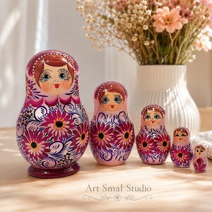 Collection Zoia de 5 poupées russes peintes à la main dans des tons violet framboise nacrés, mise en scène sur une table en bois près d'un vase fleuri. Une décoration artisanale inspirée du folklore russe, pleine de charme et de douceur.