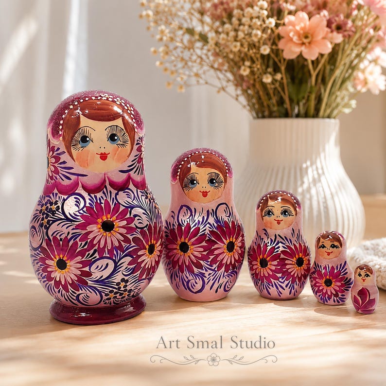 Collection Zoia de 5 poupées russes peintes à la main dans des tons violet framboise nacrés, mise en scène sur une table en bois près d'un vase fleuri. Une décoration artisanale inspirée du folklore russe, pleine de charme et de douceur.