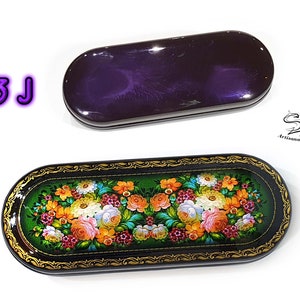 Puede incluir: Una caja ovalada de color negro y dorado con un dise&ntilde;o floral pintado en la tapa. La caja est&aacute; cerrada y el dise&ntilde;o floral es visible. La caja est&aacute; sobre un fondo blanco.