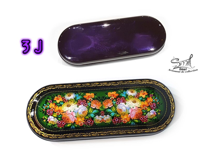 Puede incluir: Una caja ovalada de color negro y dorado con un dise&ntilde;o floral pintado en la tapa. La caja est&aacute; cerrada y el dise&ntilde;o floral es visible. La caja est&aacute; sobre un fondo blanco.