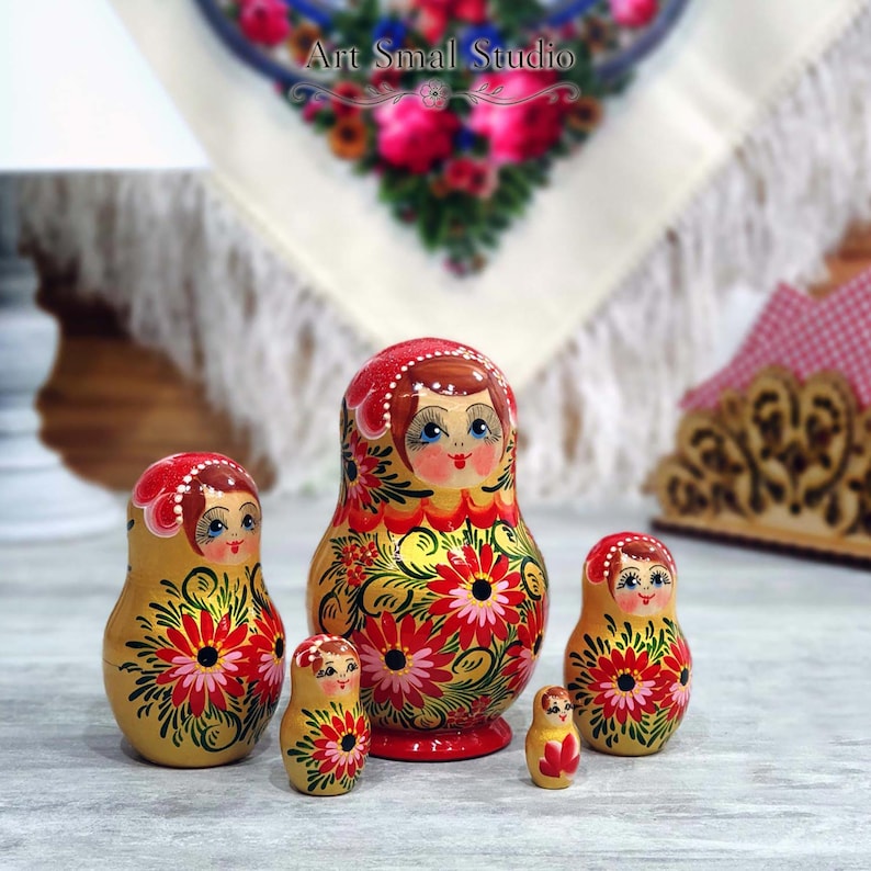 Ensemble complet de 5 matriochkas russes jaune doré, peintes à la main avec de grands motifs floraux rouges et roses. Présentation dans une ambiance chaleureuse inspirée des traditions russes, idéale pour la décoration et les collectionneurs.