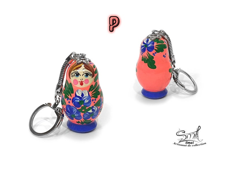 May include: Two Russian nesting doll keychains. The dolls are painted with a coral background, blue floral designs, and a blue base. Each keychain has a silver-colored metal ring and chain. The dolls have a traditional painted face.
