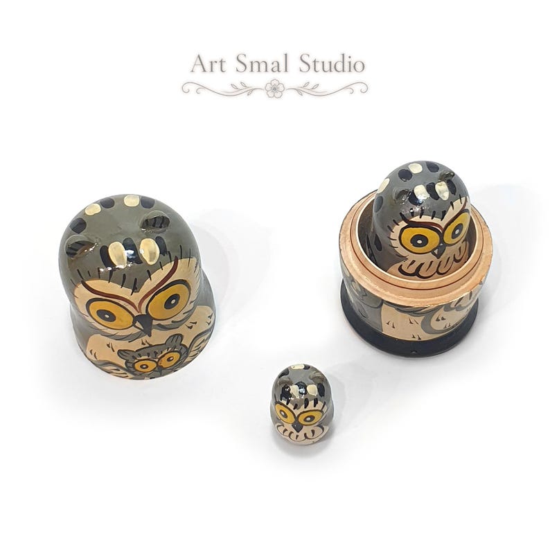 Top view of an owl-themed three-piece Russian nesting doll set, showing the hand-painted gray and cream designs, golden-yellow eyes and detailed owl motifs, with one doll opened to reveal a smaller nesting figure on a white background
