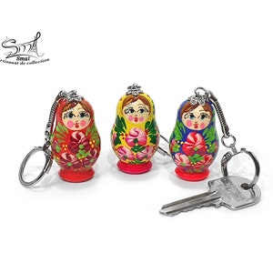 May include: Three hand-painted Russian nesting doll keychains. Each doll features a unique floral design in red, yellow, and blue, with a silver keychain attached. A silver key rests in front of the dolls. The dolls have a red, yellow, and blue base.