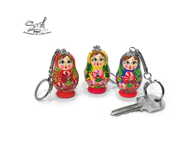 May include: Three hand-painted Russian nesting doll keychains. Each doll features a unique floral design in red, yellow, and blue, with a silver keychain attached. A silver key rests in front of the dolls. The dolls have a red, yellow, and blue base.