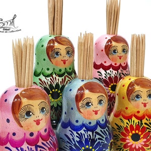 Puede incluir: Cinco muñecas rusas de madera de colores, cada una con una cara pintada y un diseño floral, se utilizan como porta palillos. Los palillos sobresalen de la parte superior de cada muñeca.