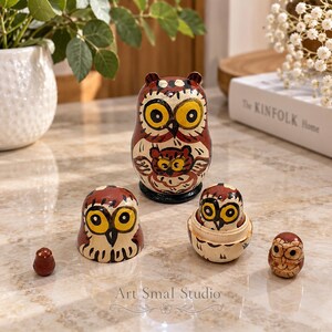 Owl matryoshka nesting doll set, five hand-painted wooden owls in burgundy, cream and gold, including a remarkably tiny miniature owl. Displayed on a polished marble table in a warm interior with green plants, white flowers and a book in the background