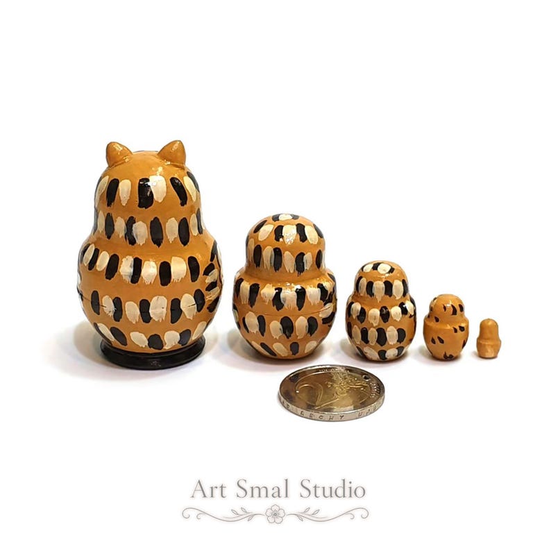 Shown from the back, five graduated owl nesting dolls reveal their rounded wooden forms and decorative black, cream and golden-brown pattern, with the smallest inner pieces beside a euro coin for scale; a miniature Russian matryoshka set on a clean white background