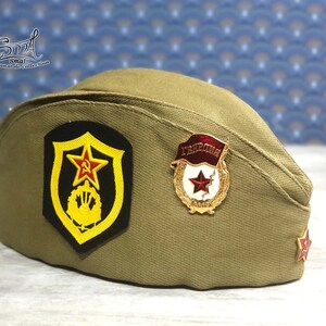 Może przedstawiać: Czapka w wojskowym stylu w kolorze khaki z czerwoną gwiazdą i żółtym emblematem z przodu. Czapka ma również czerwoną gwiazdę i złoty emblemat z napisem "Україна".