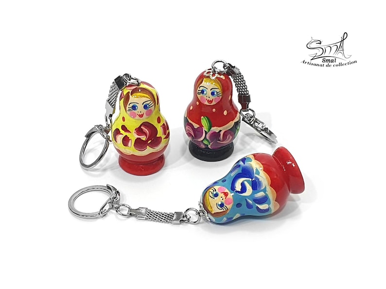 May include: Three colorful wooden nesting doll keychain charms. The dolls are painted with traditional Russian designs and have silver keychains.