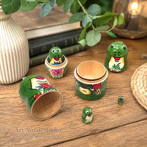 An artistic close-up shows the five-piece frog nesting doll collection displayed open on a rustic wooden table, surrounded by leafy plants and warm home décor
