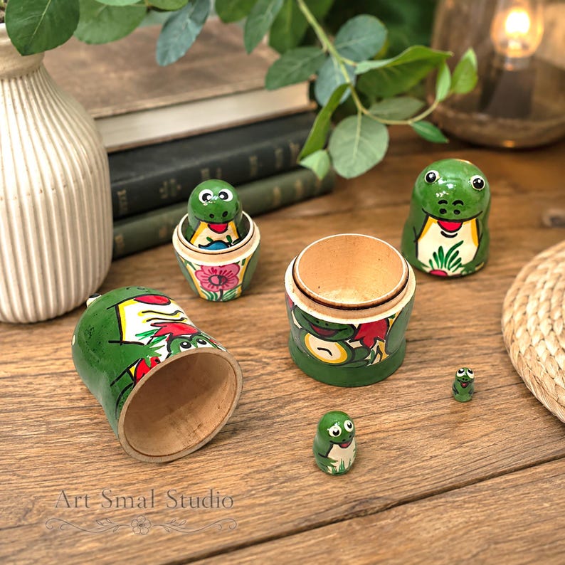 An artistic close-up shows the five-piece frog nesting doll collection displayed open on a rustic wooden table, surrounded by leafy plants and warm home décor