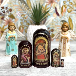 May include: A collection of Russian nesting dolls, or Matryoshka dolls, featuring religious imagery. The dolls are various sizes and colors, with the largest doll depicting the Virgin Mary and baby Jesus. Two angel figurines flank the dolls.