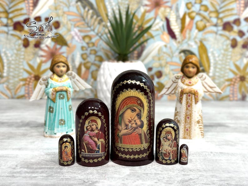May include: A collection of Russian nesting dolls, or Matryoshka dolls, featuring religious imagery. The dolls are various sizes and colors, with the largest doll depicting the Virgin Mary and baby Jesus. Two angel figurines flank the dolls.