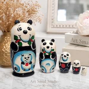Panda matryoshka nesting dolls displayed as a five-piece Russian wooden doll set, with hand-painted black, white, blue, green and red details. The graduated dolls are arranged on a marble surface in an elegant home setting with dried flowers and soft natural light