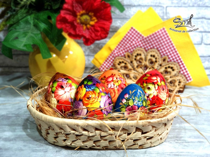 Cinq œufs de Pâques en bois peints à la main style russe dans un panier en osier. Les œufs sont décorés de motifs floraux colorés et sont disposés dans un nid de paille.
