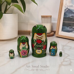 Set against a bright marble tabletop, five green frog nesting dolls are displayed in a modern home interior with plants, a candle and framed artwork in the background