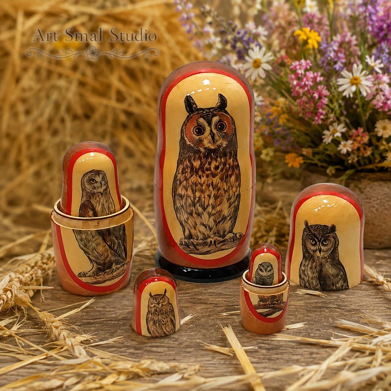 Owls hand-painted on a set of five Russian nesting dolls, featuring different owl species and detailed plumage. The Matryoshkas are arranged on a rustic wooden table surrounded by straw and dried wheat, with a colorful bouquet of wildflowers in the background, creating a warm countryside atmosphere