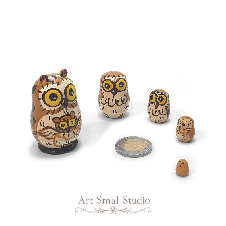 A charming family of five owl figures with golden eyes, painted in warm brown and cream tones, forms a miniature Russian nesting doll set, with the tiny inner pieces displayed beside a euro coin for scale against a clean white background