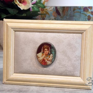 May include: A framed oval portrait of a woman in a vintage style. The portrait is set against a light beige background within a light wood frame. The portrait is in a silver-coloured oval frame. The frame is rectangular.