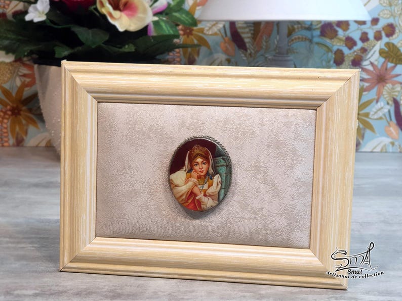 May include: A framed oval portrait of a woman in a vintage style. The portrait is set against a light beige background within a light wood frame. The portrait is in a silver-coloured oval frame. The frame is rectangular.